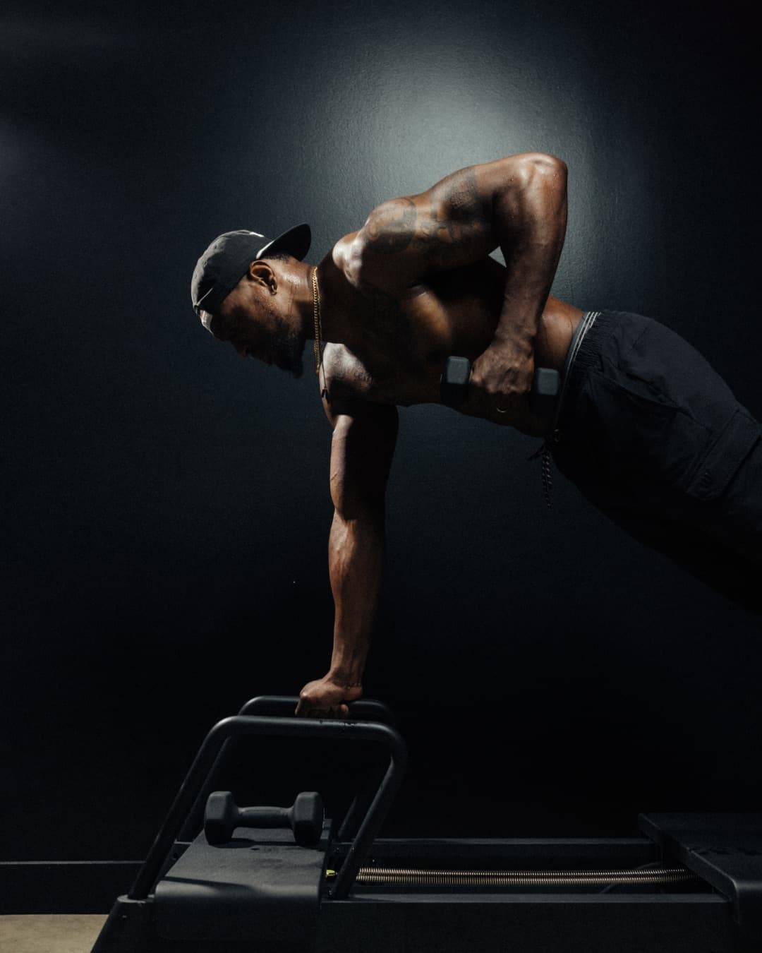 A member holding a loaded plank on the reformer, dumbbell rowed to the ribs, arms and back under visible tension.